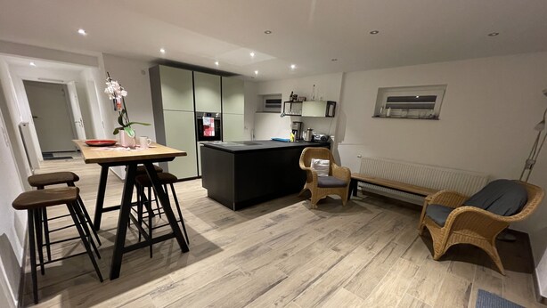 A modern kitchen with a central island, bar stools, a wicker chair, and a bench. - Kellerwohnung (Seelbach)