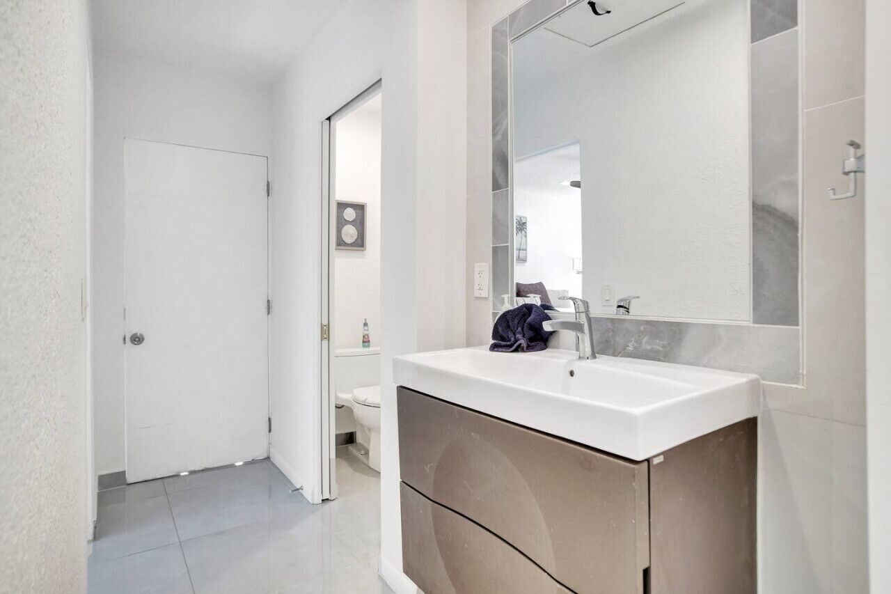Een moderne badkamer met een wit wastafel, een grote spiegel en een deur naar een andere kamer.