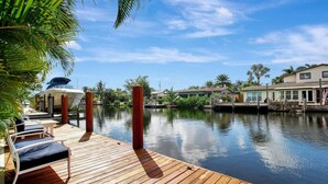 A wooden deck with lounge chairs and a boat docked by a calm water body, surrounded by palm trees and buildings. - Wilton on the Water -Courtyard & Heated Pool 4/2.5 (Wilton Manors)