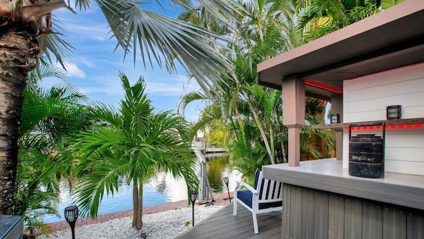 A deck with a white chair and a small table, surrounded by palm trees and a body of water. - Wilton on the Water -Courtyard  & Heated Pool 4/2.5 (Wilton Manors)