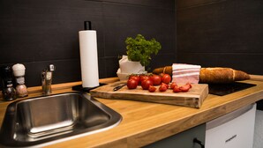 Una encimera de cocina con un lavamanos, una tabla de cortar con tomates y una barra de pan, un rollo de servilletas de papel y una planta en maceta.