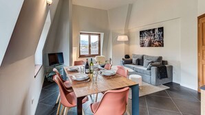A dining area with a wooden table, pink chairs, and a television. There is a sofa and a painting on the wall.