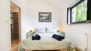 A bedroom with a bed, two bedside tables, a radiator, a window with a view of greenery, and a door leading to another room. - The Cabin in the Tamar Valley Cornwall (Gunnislake)