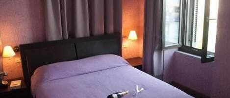 A neatly made bed with a purple bedspread, two bedside lamps, and a window with curtains.
