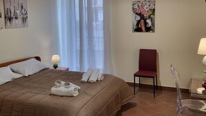 Une chambre d’hôtel avec un lit, une chaise, une table avec une lampe, un tableau et une fenêtre avec des rideaux.