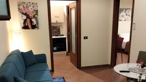 Un salon avec un canapé, une table basse et un tapis. Au fond, on aperçoit une cuisine avec une cuisinière et des placards.