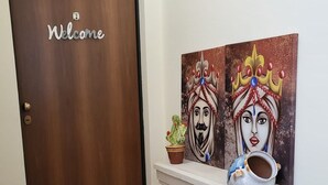Une porte en bois avec une pancarte « Bienvenue », une console blanche ornée d’un vase et d’une plante en pot, et deux tableaux colorés accrochés au mur.
