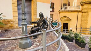 Une statue représentant un homme assis, sur une rue pavée de pierres, avec des bâtiments en arrière-plan.