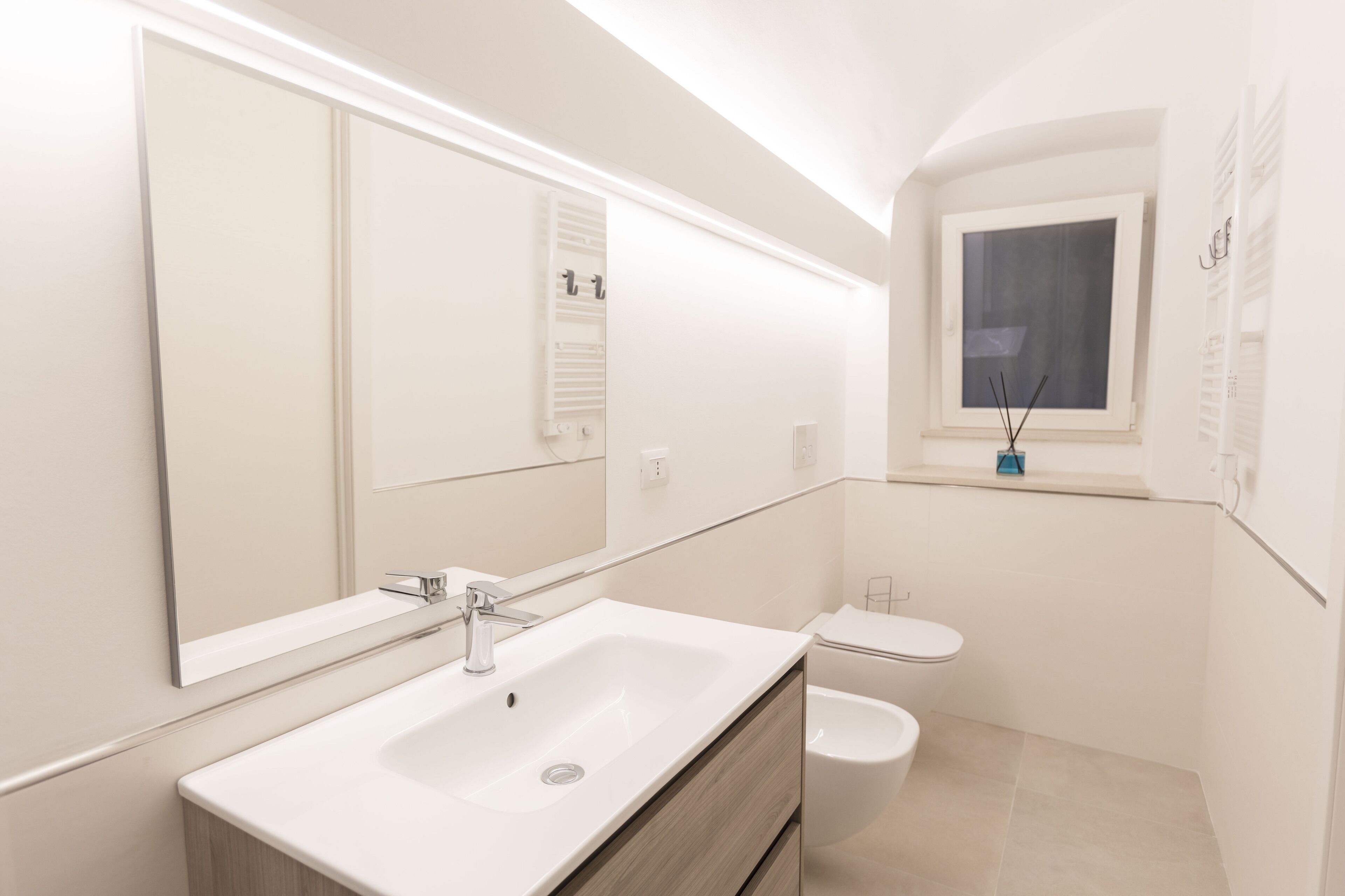 Un baño moderno con un espejo grande, un lavamanos blanco, un inodoro y una ventana con vistas.