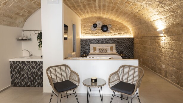 A compact room with a stone ceiling, a bed with patterned bedding, a small kitchen area, and two wicker chairs. - Torre Canina Suite & Rooms  (Trani)