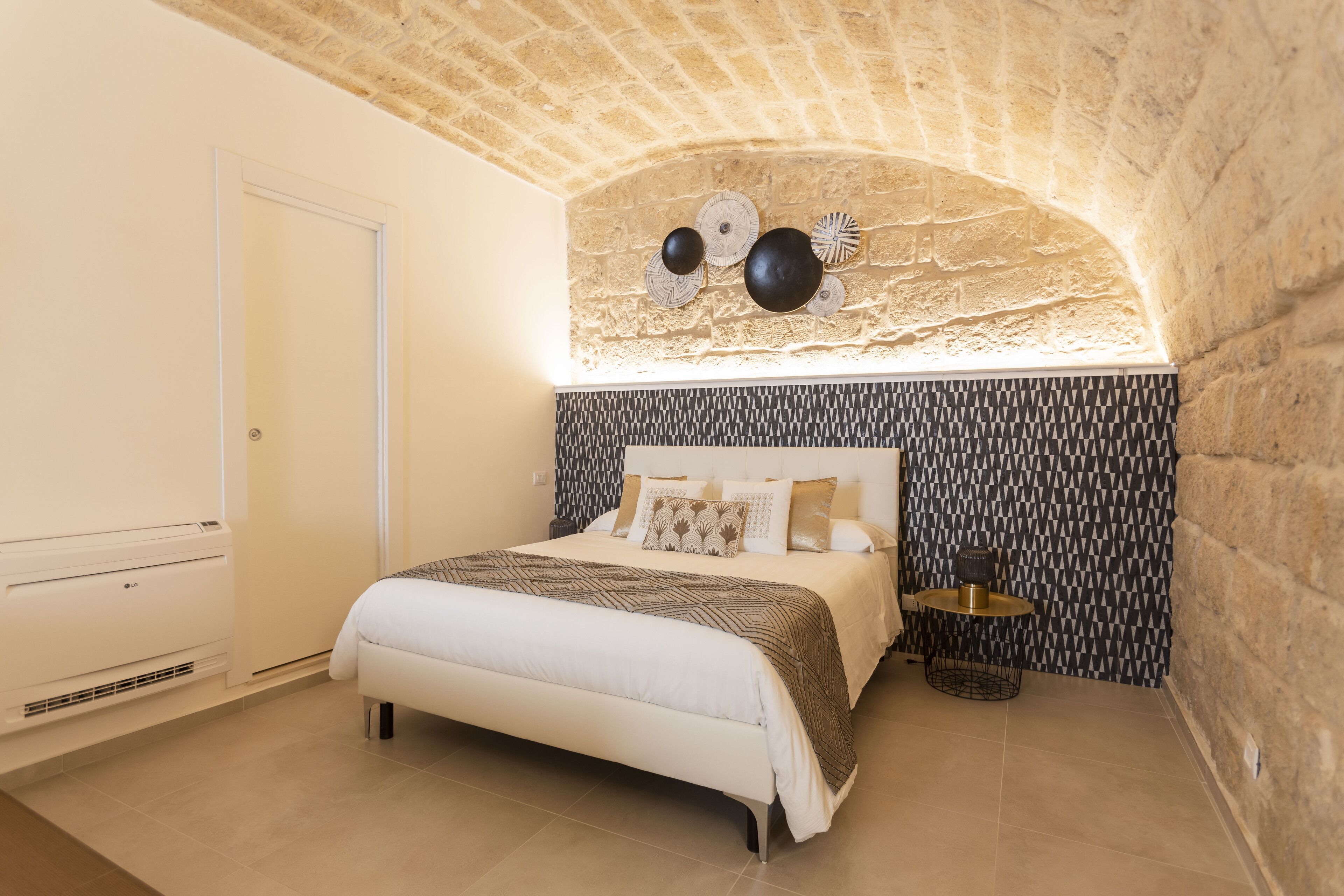 Ein Schlafzimmer mit einer Decke aus Steinbögen, einem Bett mit gemusterten Textilien, einem Nachttisch mit Lampe und einer Wand mit Muster.