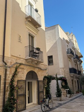 A building with a sign indicating it is a hotel, featuring a balcony with a chair and a potted plant, a bicycle parked in front, and a stone facade. - Torre Canina Suite & Rooms (Trani)