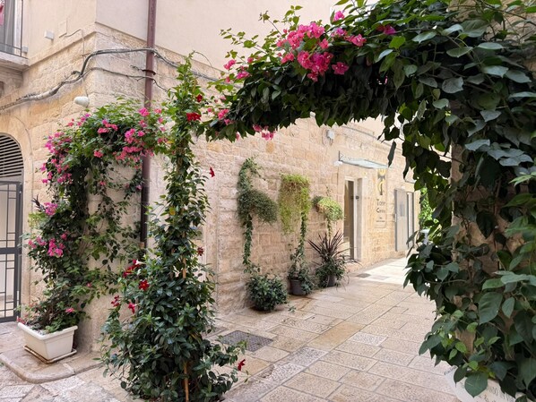 Un estrecho callejón adoquinado con un edificio a un lado, que presenta un arco cubierto de hiedra y flores rosadas.