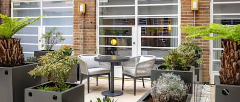 An outdoor seating area with a round table and chairs, surrounded by square planters with various plants.