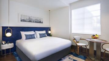 A modern hotel room with a large bed, bedside tables, a desk with chairs, and a large window with blinds.