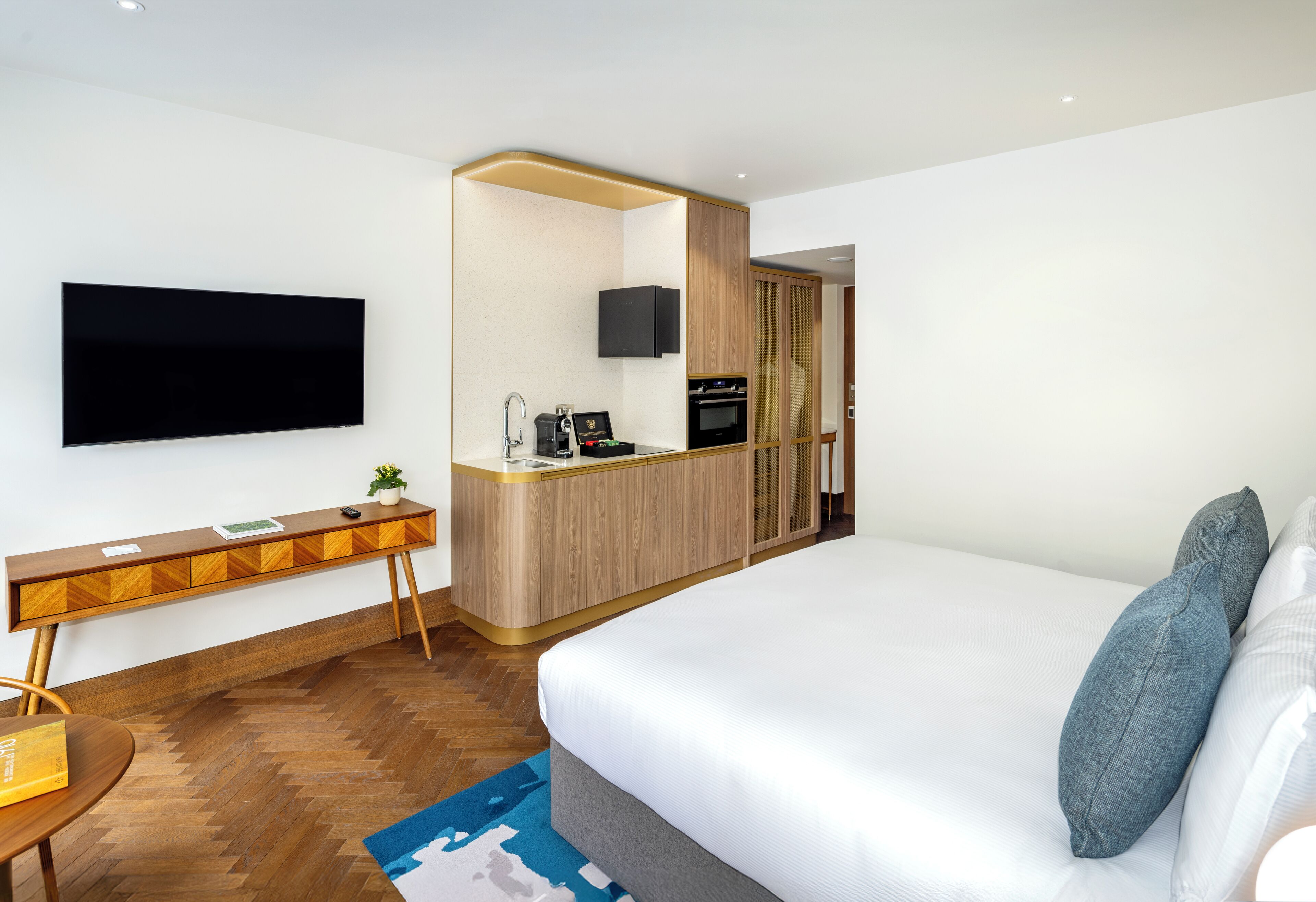 Une chambre d’hôtel moderne dotée d’un grand lit, d’un bureau avec un téléviseur à écran plat, d’une kitchenette et d’une salle de bain visible à travers une porte ouverte.