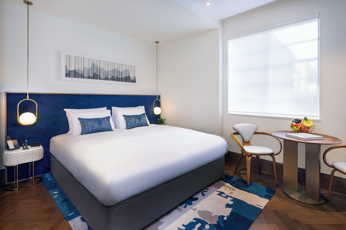 A modern hotel room with a large bed, bedside tables, a desk with chairs, and a large window with blinds.