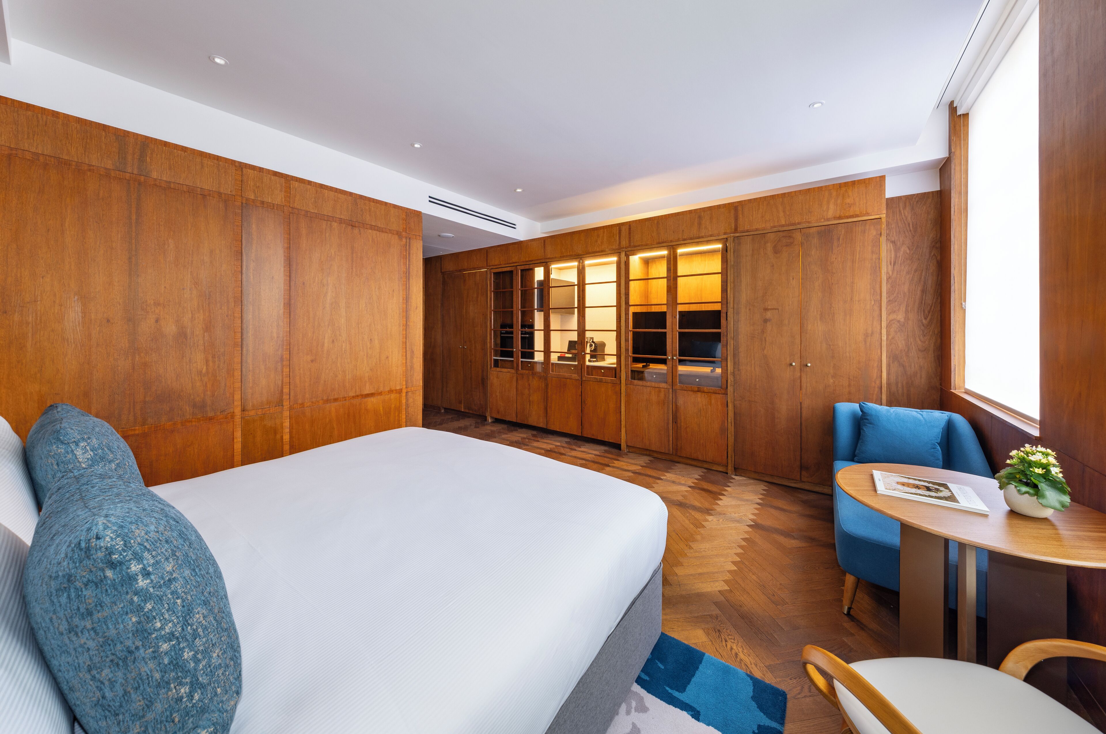 A modern hotel room with a large bed, wooden wardrobe, a small table with a plant, and a blue chair.