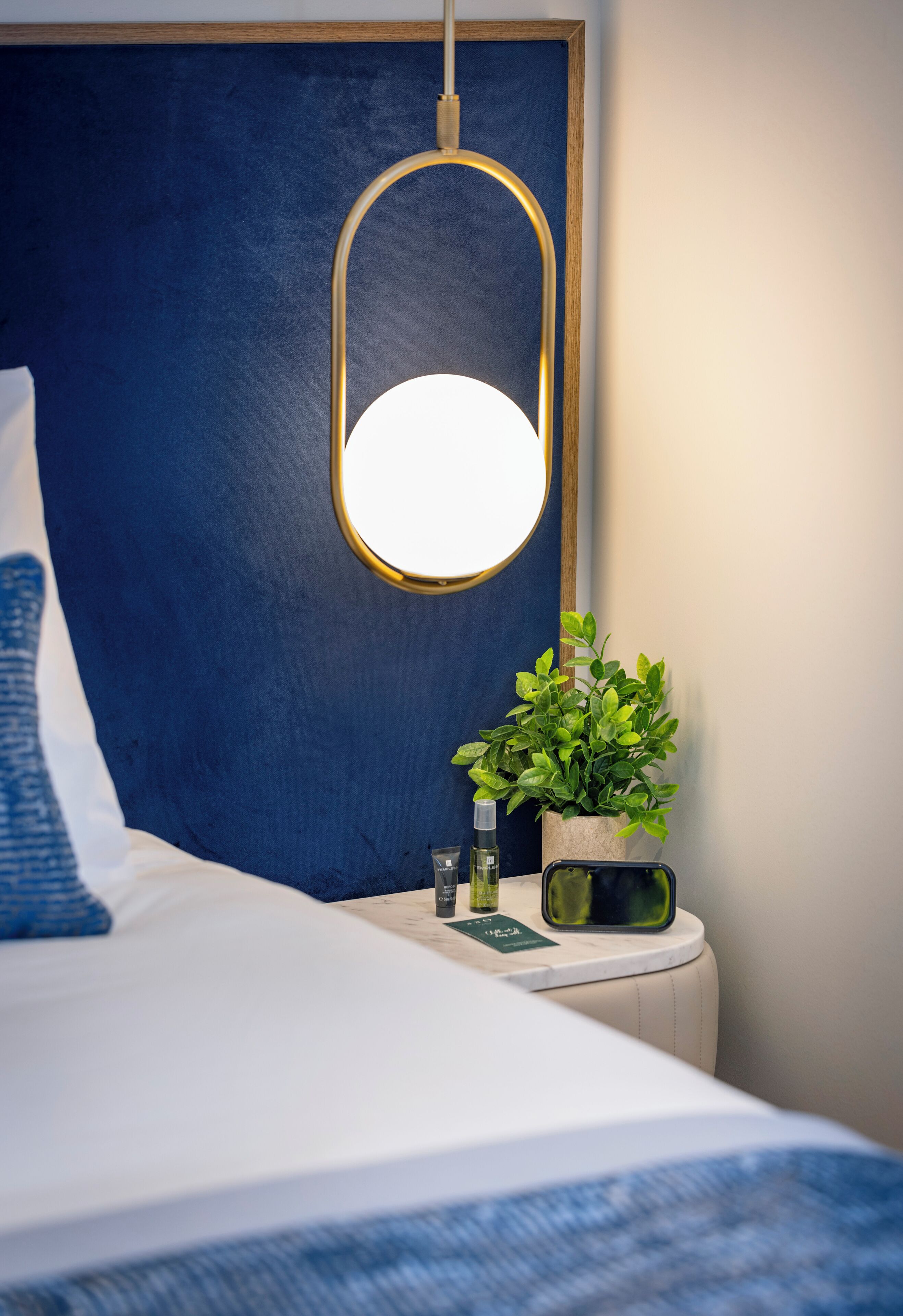 Een net opgemaakt bed met een blauw hoofdbord, een nachtkastje met een groene plant, een kleine lamp en een smartphone.