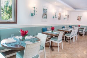 A restaurant with round tables, white wicker chairs, teal cushions, and artwork on the walls. - The Phoenix Resort (San Pedro)