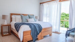 A bedroom with a wooden bed, white bedding, a blue throw blanket, two bedside tables with lamps, and a view of greenery through a balcony door.