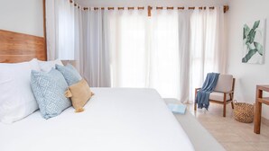 A neatly made bed with patterned pillows, a wooden headboard, a chair with a blanket, and a woven basket on a tiled floor.