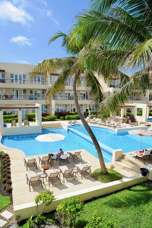 A pool area with lounge chairs and umbrellas, surrounded by palm trees and a building with balconies. - The Phoenix Resort (San Pedro)