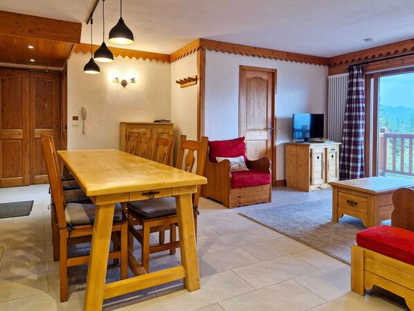 A wooden dining table with chairs, a sofa, a television, and a wooden cabinet in a room with a wooden ceiling and walls.