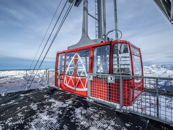 Um funicular vermelho com um logotipo triangular, suspenso por cabos, com vista para uma paisagem montanhosa coberta de neve.