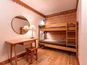 A bunk bed with a wooden frame and a single bed below. There is a small wooden desk with a chair and a lamp. A round mirror hangs on the wall above the desk.