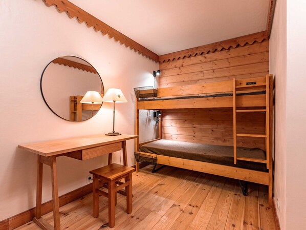 A bunk bed with a wooden frame and a single bed below. There is a small wooden desk with a chair and a lamp. A round mirror hangs on the wall above the desk.