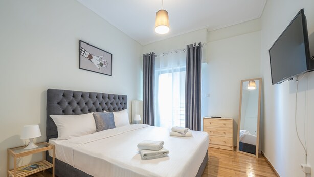 A neatly made bed with a grey headboard, a bedside table with a lamp, a wooden dresser, and a large mirror on the wall. - Athens Apartment 5-min to Metaxourgeio Metro  (Athina)