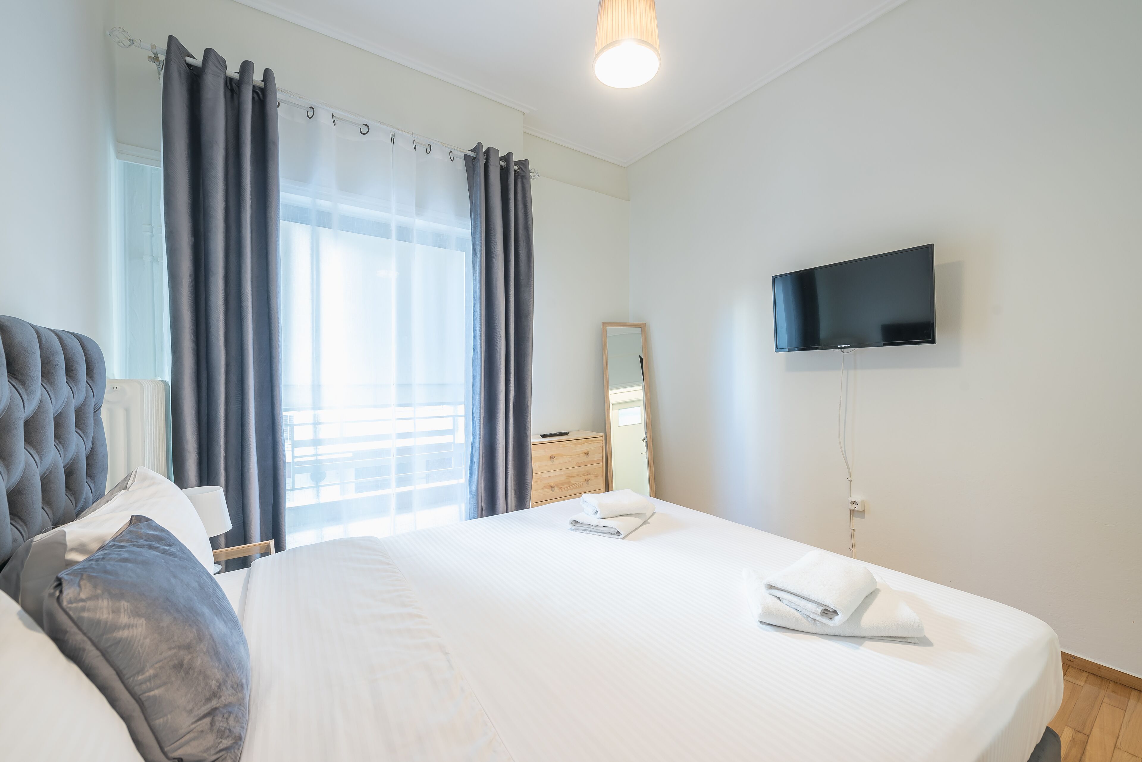 A neatly made bed with pillows, a bedside table, a flat-screen TV, and a window with curtains.