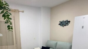 Ein kompakter Wohnbereich mit einer Couch, zwei kleinen runden Tischen, einem weißen Kühlschrank und einer Pflanze.