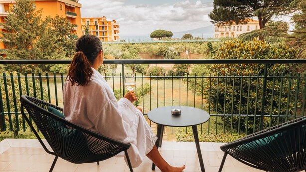 A balcony with two chairs and a table, overlooking a garden and buildings. - Sama Apartments – Elegance, Comfort and Sea View in the Heart of Sorrento (Sorrento)