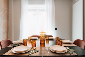 Una mesa de comedor dispuesta con platos, cubiertos y cristalería decorada, con una ventana y cortinas al fondo.