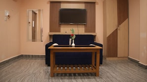 A room with a dark blue sofa, a wooden table with a vase of flowers, a flat-screen TV mounted above the sofa, a wardrobe, and a mirror on the wall.