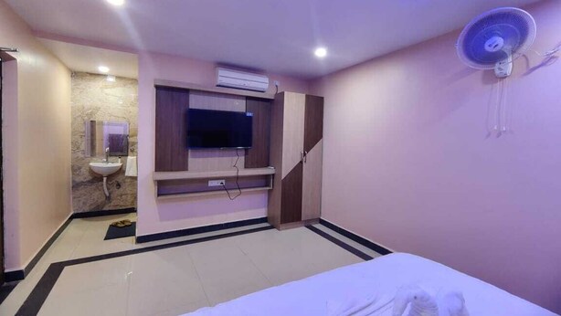 A hotel room with a bed, a TV mounted on the wall, a ceiling fan, and a small bathroom with a sink and mirror.