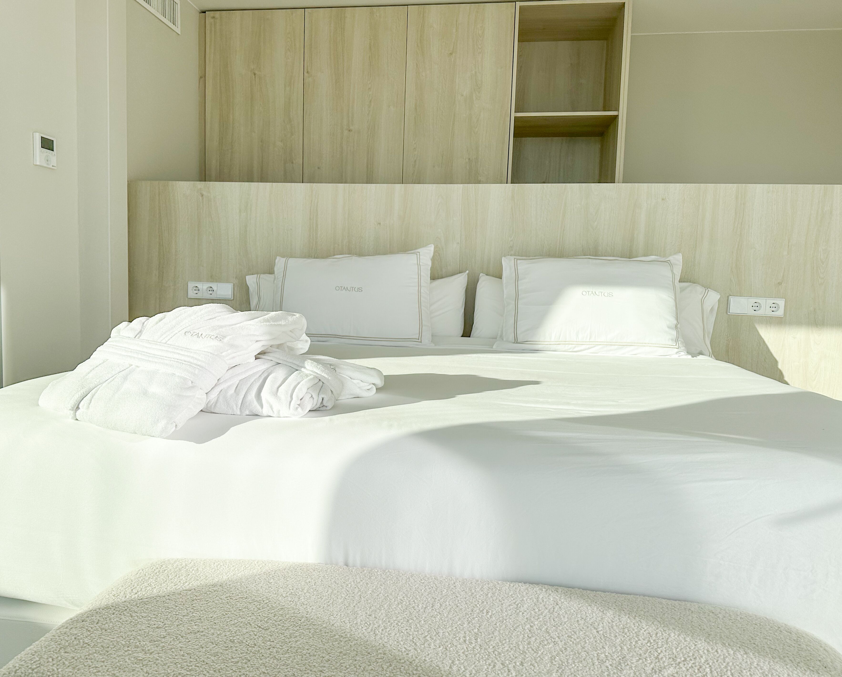 A neatly made bed with white linens and pillows, a white bathrobe on the bed, and a wooden headboard.