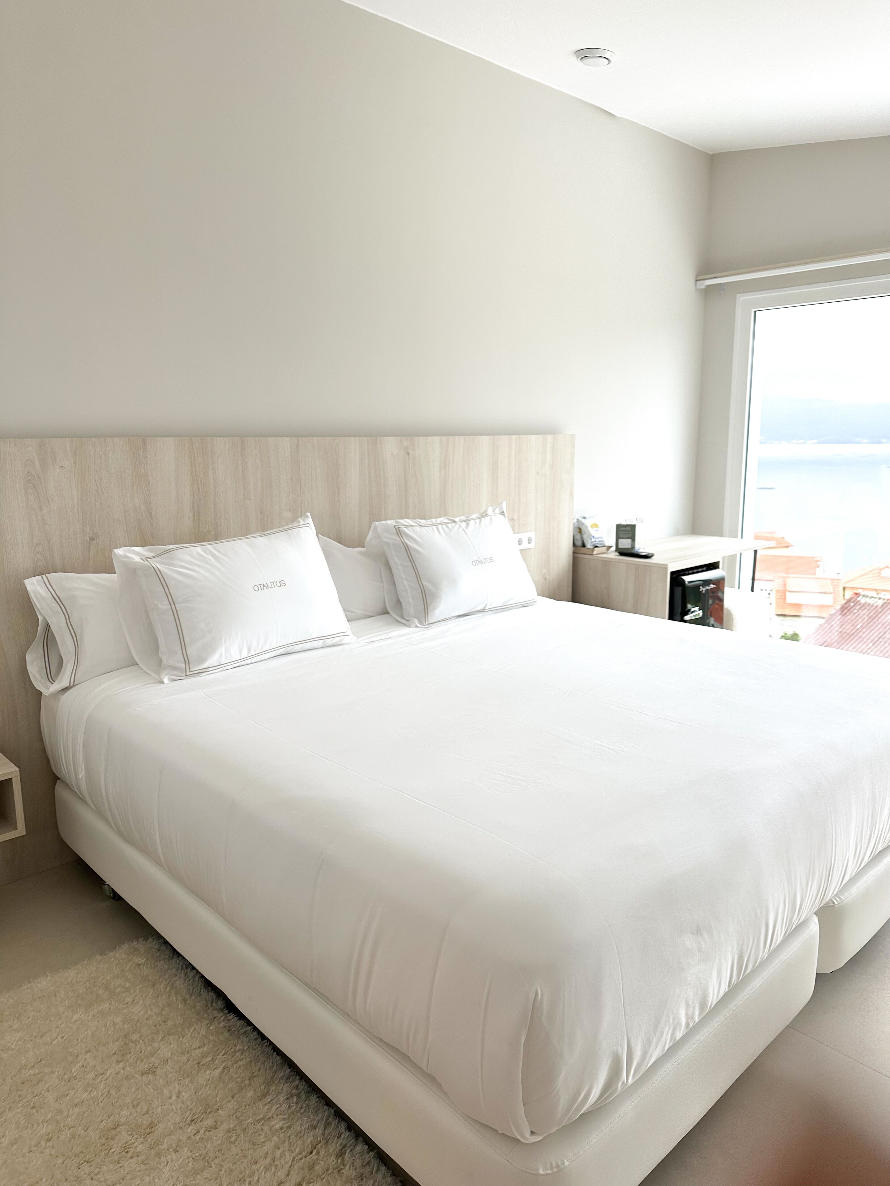 Una cama grande con ropa de cama y almohadas blancas, un cabecero de madera, una mesita de noche con una lámpara y vistas al mar a través de un ventanal amplio.