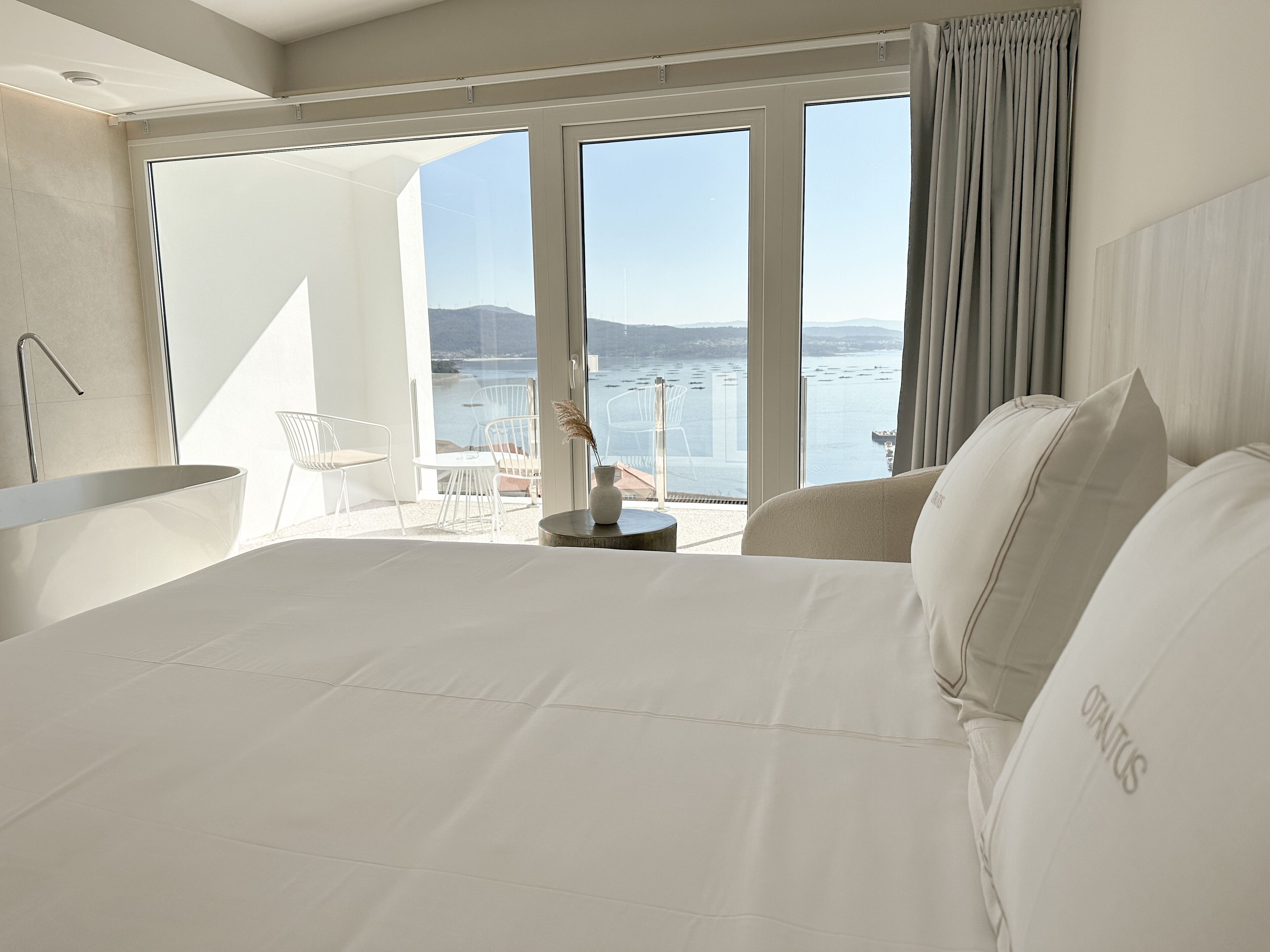 A modern hotel room with a large bed, a bathtub, and a balcony with a view of the sea.