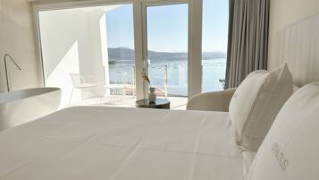 A modern hotel room with a large bed, a bathtub, and a balcony with a view of the sea.