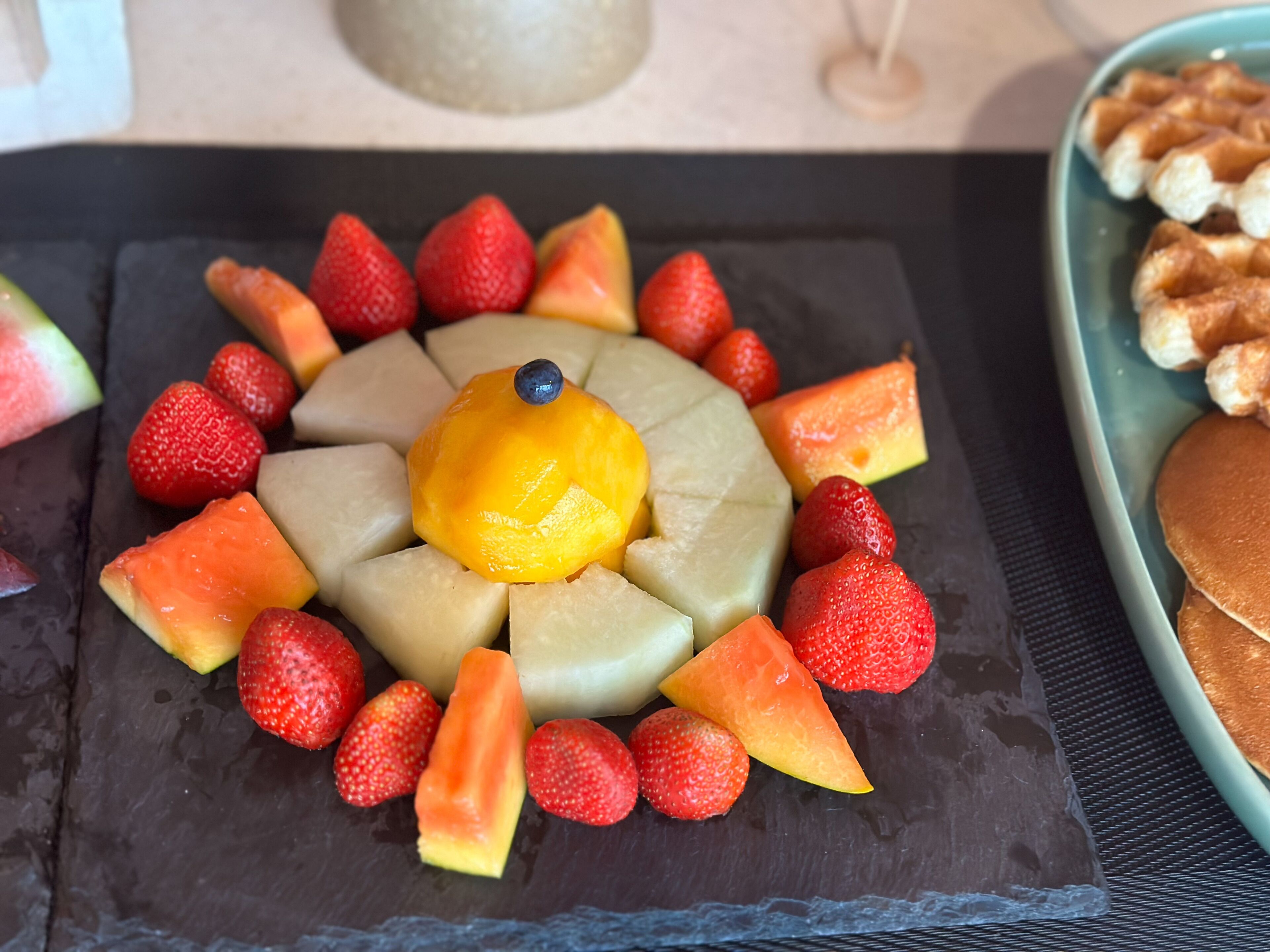 A fruit platter with strawberries, cantaloupe, and a yellow fruit in the center, arranged on a dark slate plate.