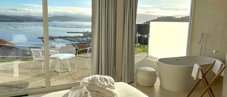 A modern bedroom with a large window, a bathtub, and a view of the sea.