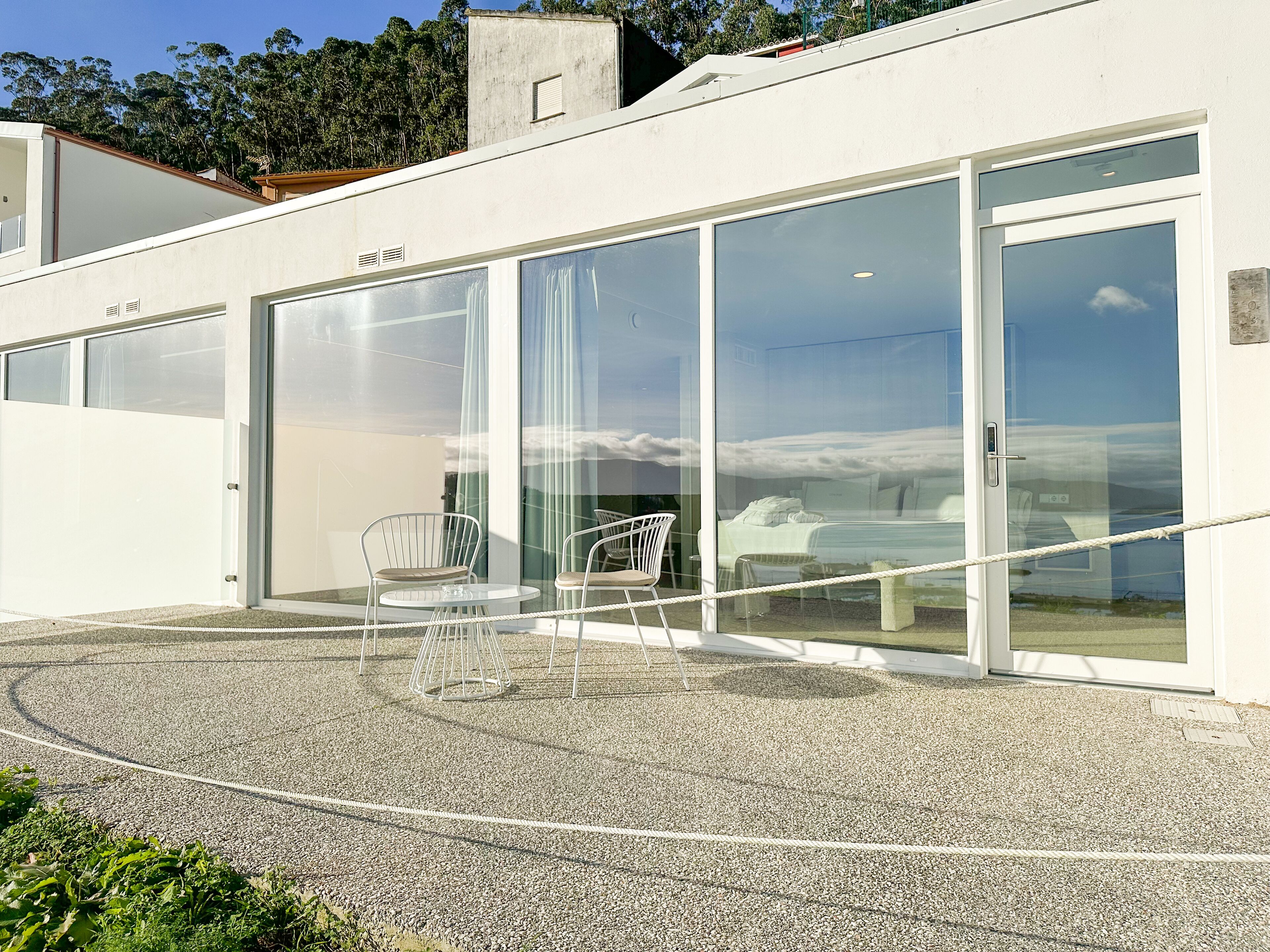 Un bâtiment moderne doté de grandes portes et fenêtres en verre, d’une terrasse en gravier avec deux chaises, et d’un lit visible à travers le verre.