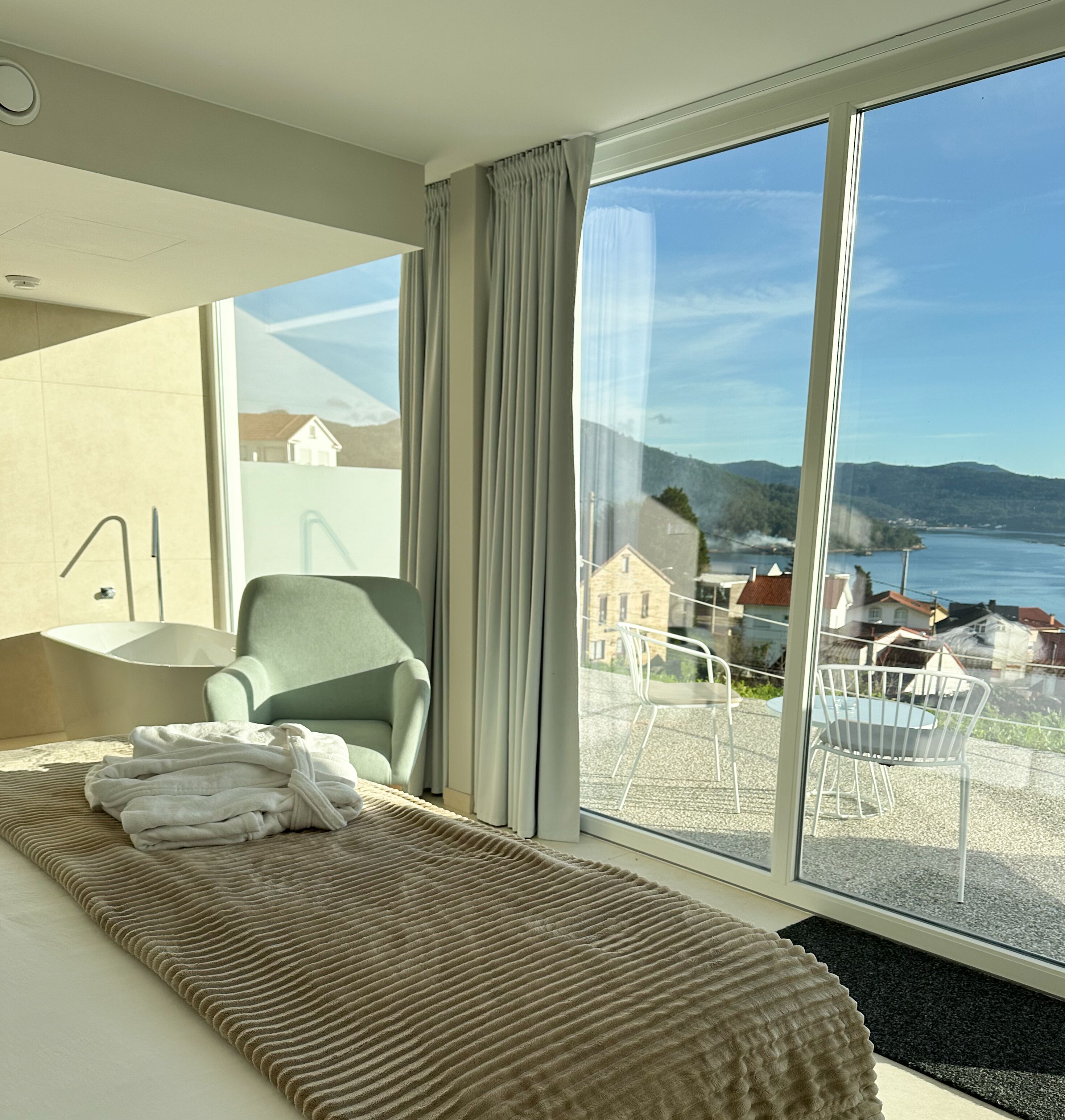 Habitación de hotel moderna con un ventanal grande, una cama con manta color beige, una silla y vistas a un cuerpo de agua y edificios.