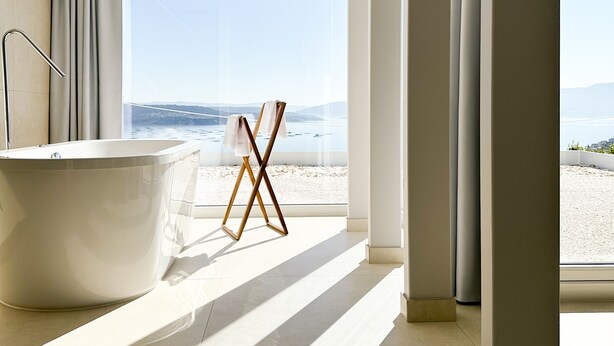 A modern bathroom with a freestanding bathtub, a wooden drying rack, and a large window offering a view of the beach. - Otantus Hotel (Muros)