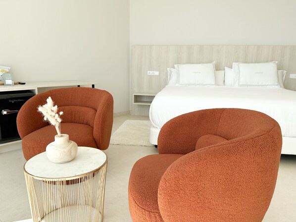 Une chambre d’hôtel moderne avec un grand lit, deux fauteuils confortables, une petite table ronde et un vase contenant des fleurs séchées.