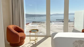 Habitación de hotel con un ventanal amplio que ofrece vistas a una playa y al agua. Se observa una cama, una silla y una mesita.