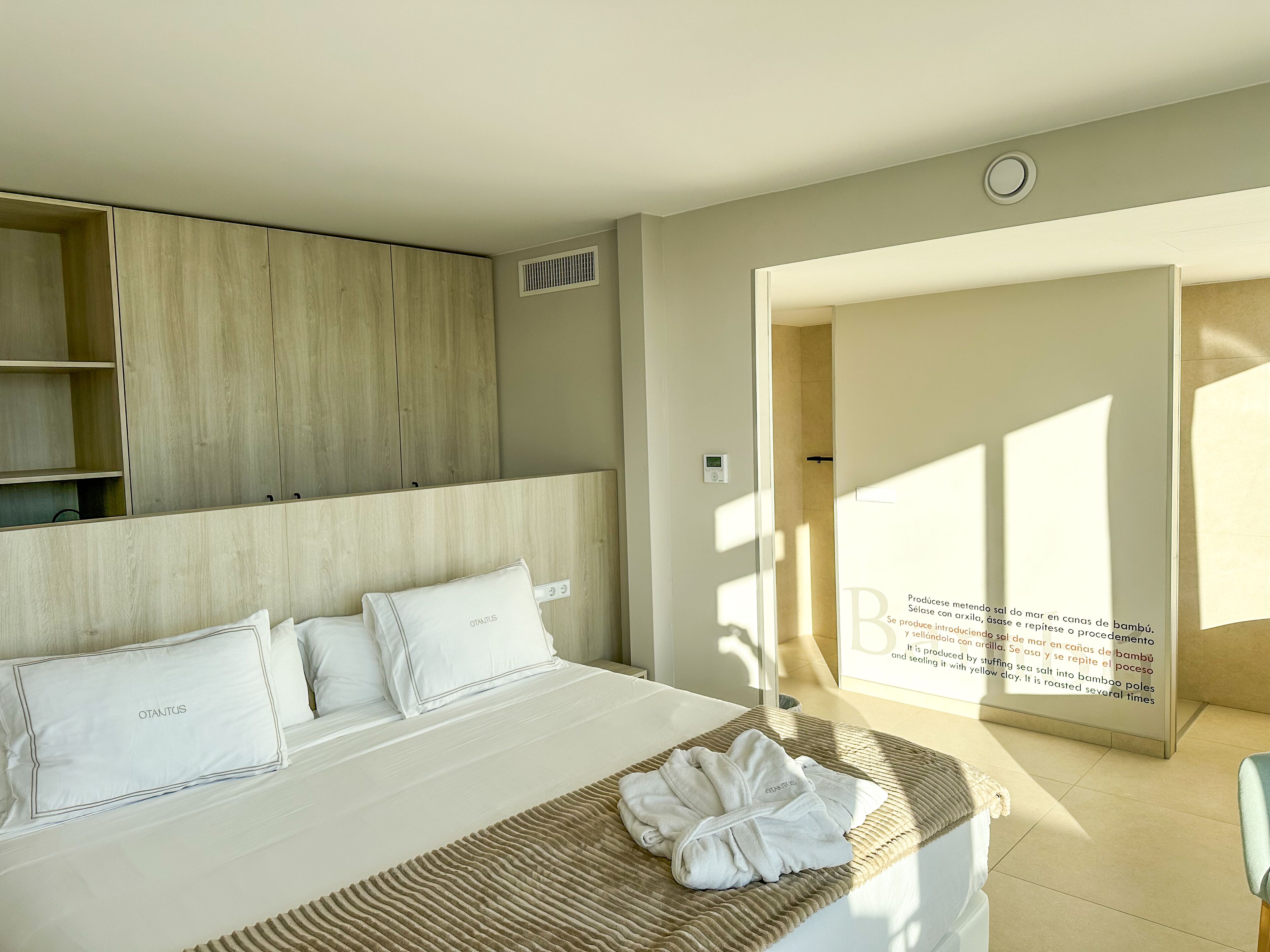A hotel room with a bed, a wooden headboard, a wall-mounted air vent, and a door leading to another room.